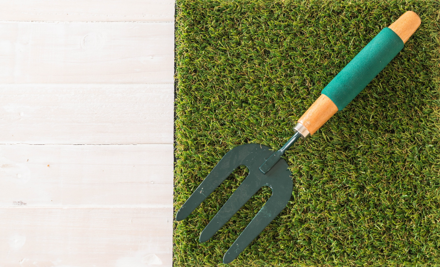 Un rastrillo de jardinería con mango de madera y agarre de goma verde colocado sobre césped artificial. La imagen destaca la herramienta y una sección de césped sintético, representando el mantenimiento y cuidado del jardín.